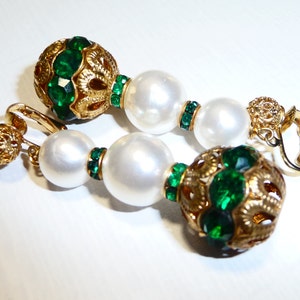 May include: A pair of gold tone clip-on earrings with green and white beads. The earrings feature a large round green bead with gold filigree and a smaller round white bead.