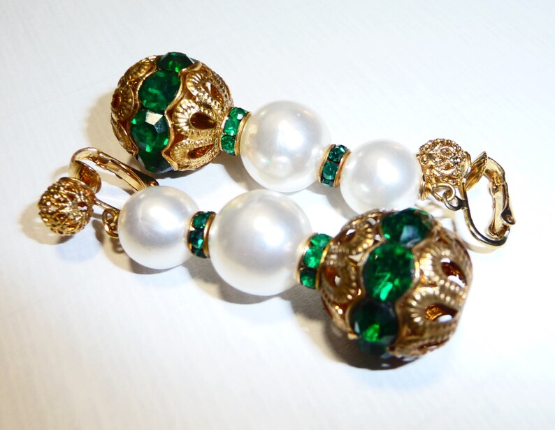 May include: A pair of gold tone clip-on earrings with green and white beads. The earrings feature a large round green bead with gold filigree and a smaller round white bead.