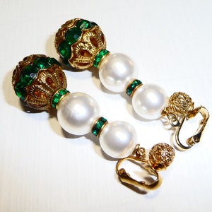 May include: A pair of gold tone clip-on earrings with green and white beads. The earrings feature a large round bead with green rhinestones and a gold filigree design. The beads are strung on a gold chain with smaller green rhinestones.