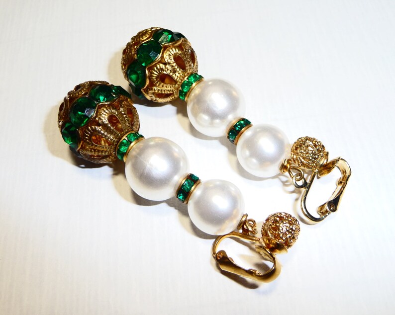 May include: A pair of gold tone clip-on earrings with green and white beads. The earrings feature a large round bead with green rhinestones and a gold filigree design. The beads are strung on a gold chain with smaller green rhinestones.