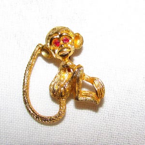 May include: A gold-colored monkey brooch with red gemstone eyes. The monkey is in a sitting position with its tail curled. The brooch has a textured surface and is set against a white background. A vintage-style jewelry piece.