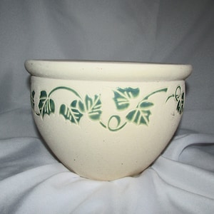 May include: A white ceramic planter with a green ivy design. The planter is round and has a wide opening.