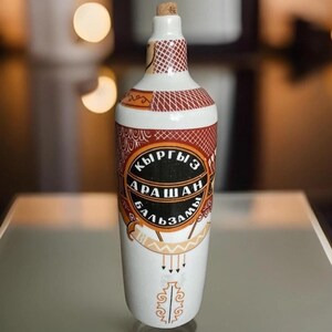 May include: A white ceramic bottle with a cork stopper. The bottle has a red and brown patterned design around the neck and a label with Cyrillic text. The label is black with gold accents. The bottle is likely for a beverage.