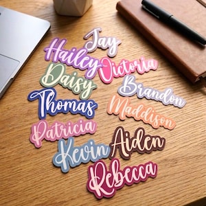 May include: A collection of die-cut stickers featuring various names in cursive script, including Jay, Hailey, Victoria, Daisy, Brandon, Thomas, Maddison, Patricia, Kevin, Aiden, and Rebecca, each rendered in distinct color palettes with a white border.