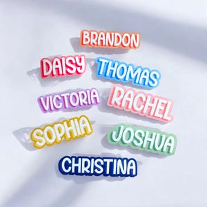 May include: A collection of eight custom name tags featuring the names Brandon, Daisy, Thomas, Victoria, Rachel, Sophia, Joshua, and Christina. Each name is rendered in a bold, white, sans-serif font with a colourful, layered, three-dimensional shadow effect.