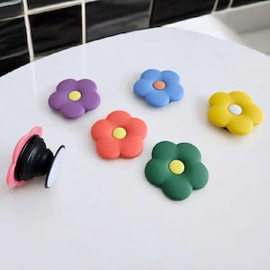 May include: A selection of flower-shaped phone grips in various colours: purple, blue, red, yellow, and green. One pink grip is extended, revealing the black base and white adhesive. These accessories add a floral touch to any phone.