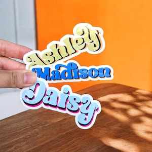 Può includere: Tre adesivi in vinile fustellati con i nomi Ashley, Madison e Daisy in un carattere audace di ispirazione retrò. L'adesivo Ashley è giallo pallido, l'adesivo Madison è blu reale e l'adesivo Daisy è azzurro con contorni rosa.