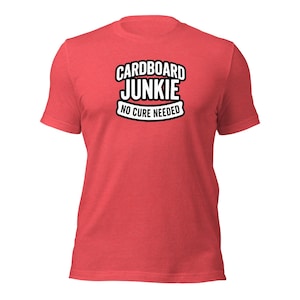 Op de afbeelding: Een rood t-shirt met korte mouwen voorzien van de tekst "CARDBOARD JUNKIE" in dikke zwarte letters met een witte omlijning, gevolgd door de tekst "NO CURE NEEDED" in een kleiner zwart lettertype binnen een witte banner op de borst.