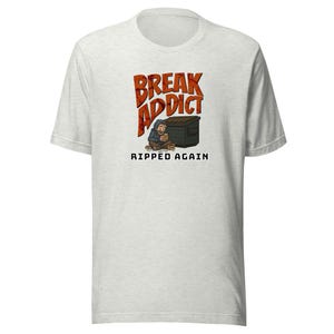 May include: A light heather gray short-sleeve t-shirt featuring a graphic of a person sitting by a black dumpster. The text reads BREAK ADDICT in large orange letters above the figure and RIPPED AGAIN in black text below the illustration.