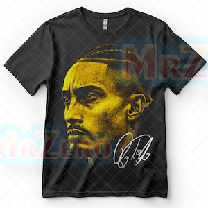 May include: Black t-shirt featuring a golden portrait of a man with a beard and braided hair. The image includes a signature in white. The shirt is a crew neck style.
