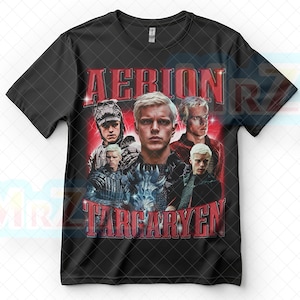 以下が含まれることがあります： 白髪の人物の画像をコラージュした黒いTシャツ。「AERION TARGARYEN」の文字が赤色のヴィンテージ風の文字でTシャツの前面にプリントされています。