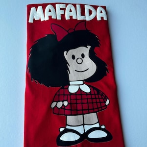 Puede incluir: Camiseta roja con una ilustración de Mafalda, personaje de cómic. El diseño incluye la cara de Mafalda, un lazo rojo y un vestido a cuadros. La palabra "MAFALDA" está impresa en blanco en la parte superior.