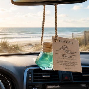 May include: A car essential oil diffuser with a teal glass bottle and wooden cap, hanging from a rearview mirror. A brown tag reads "Earthwise Sea Glass Essential Oil Car Diffuser". The background shows a beach scene.