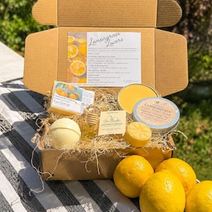 May include: A gift box labeled "Lemongrass Lovers" filled with various bath and body products. The box contains a bath bomb, soap bars, a small bottle of oil, and fresh lemons. The products are arranged on a bed of straw.