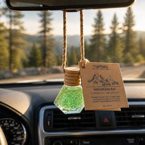 May include: A car air freshener with a clear, geometric-shaped glass bottle filled with green glitter. The bottle hangs from a woven rope and a wooden cap. A brown tag reads "MOUNTAIN AIR" and "ESSENTIAL OIL CAR DIFFUSER".