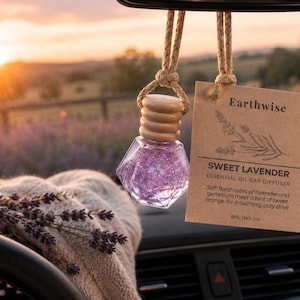 May include: A car air freshener with a clear, geometric-shaped bottle filled with purple glitter hangs from a rearview mirror. A brown tag reads "Earthwise Sweet Lavender Essential Oil Car Diffuser". A bouquet of lavender rests on the dashboard.