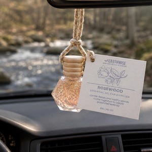 May include: A car essential oil diffuser hanging from a rearview mirror. The clear glass bottle contains gold glitter and is topped with a wooden cap. A tag reads "Rosewood Essential Oil Car Diffuser" with a floral illustration.