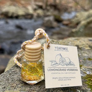 May include: A small, clear glass car diffuser bottle filled with yellow liquid and gold glitter. The bottle has a wooden cap and is attached to a beige and gold rope. A small tag reads "Lemongrass Verbena Essential Oil Car Diffuser."