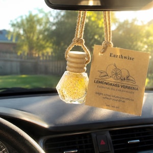 May include: A car air freshener with a glass bottle filled with yellow granules, hanging from a rearview mirror. A brown tag reads "Earthwise Lemongrass Verbena Essential Oil Car Diffuser". The car interior is visible.