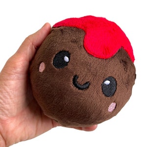 May include: A brown plush toy shaped like a chocolate pudding cup with a red chocolate topping. The toy has a smiling face with black eyes and pink cheeks.