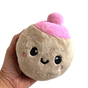 May include: A plush brown toy shaped like a round ball with a pink hat. The toy has a smiling face with black eyes and pink cheeks.