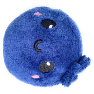 May include: A blue plush toy shaped like a whale with a smiling face. The whale has two black eyes with white highlights and a pink blush on each cheek.