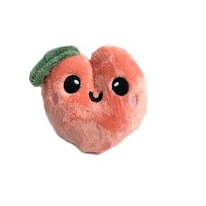 May include: A plush peach-colored heart-shaped toy with a green leaf on top. The toy has a smiling face with black eyes and a pink blush.