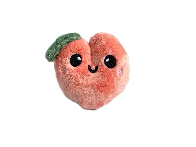 May include: A plush peach-colored heart-shaped toy with a green leaf on top. The toy has a smiling face with black eyes and a pink blush.