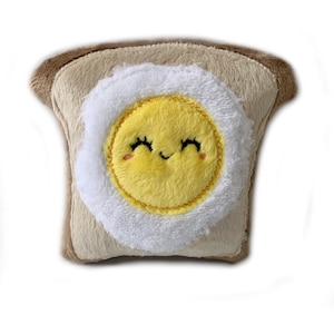 Puede incluir: Un juguete de peluche con forma de rebanada de tostada con un huevo frito encima. El huevo tiene una cara sonriente con los ojos cerrados.