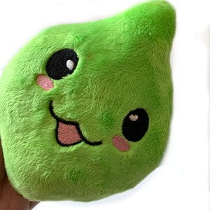 May include: A green plush toy shaped like a blob with a smiling face. The toy has black eyes, pink cheeks, and a pink tongue sticking out.