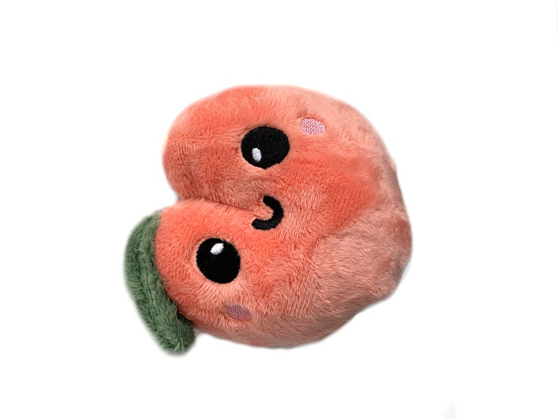May include: A plush peach-colored heart-shaped toy with a smiling face and green leaf. The toy has black eyes and a stitched-on smile.