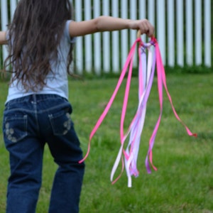 May include: A young person with long brown hair is holding a pink and purple ribbon wand. The wand is made of a wooden stick with ribbons tied to it. The person is wearing blue jeans and a gray shirt. They are standing in a grassy area with a white picket fence in the background.