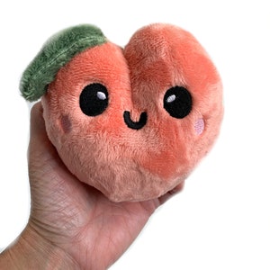 May include: A plush peach-colored peach with a green leaf on top. The peach has a smiling face with black eyes and a pink blush on its cheeks.