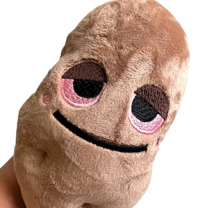 May include: A brown plush toy shaped like a potato with a smiling face. The potato has pink eyelids and black pupils, and a black stitched smile.