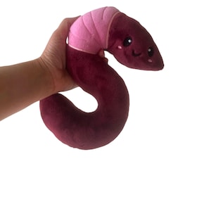 May include: A plush worm toy with a pink and burgundy color scheme. The worm has a smiling face with black eyes and a pink mouth.