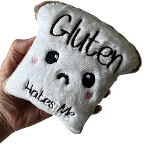 May include: A white plush bread slice with brown edging, featuring the embroidered text "Gluten Hates Me." The bread has black eyes, a curved mouth, and pink blush marks, giving it a sad expression.