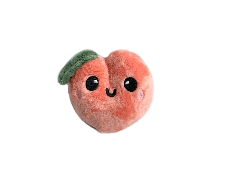 May include: A plush peach-colored peach with a green leaf and a smiling face. The peach has black eyes and a pink blush.