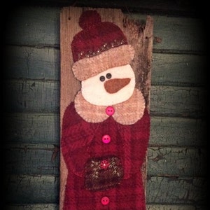 May include: A felt snowman ornament with a red coat, brown hat, and white face. The snowman is holding a small brown package and is decorated with pink buttons and green vines.
