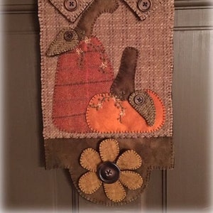 May include: A brown felt wall hanging with two pumpkins and a brown flower. The pumpkins are orange and red with brown stems. The flower has a brown center and five petals.