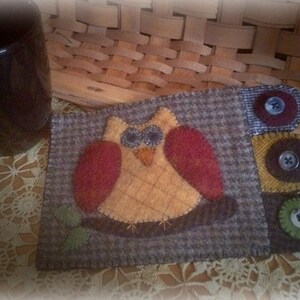 May include: A gray and brown felt owl mug rug with a yellow owl and red wings. The owl has black eyes and a brown beak. The mug rug has a brown border and a gray and white checkered pattern. There are three brown felt circles with buttons in the upper right corner.