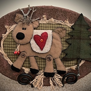 May include: A handcrafted Christmas decoration featuring a reindeer with a red heart and a green tree on an oval, brown background. The reindeer is made of felt and has a black nose, antlers, and a small bell. The tree is made of green plaid fabric.