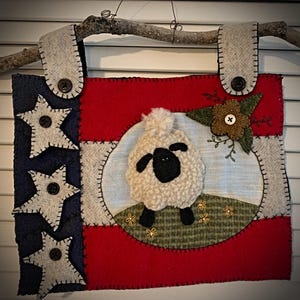 May include: A patriotic, handmade wall hanging featuring a sheep. The design incorporates red, white, and blue felt, with star and flag elements. A fluffy sheep is centered on a light blue background, with a brown flower and green leaves.