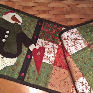 May include: A quilted table runner with a snowman holding a heart, featuring a patchwork design with red, green, and white fabrics. The runner has a brown border and is decorated with buttons.