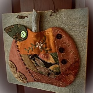 May include: A patchwork pumpkin made of fabric scraps with a twig stem and black buttons. The pumpkin is mounted on a rustic wooden board.