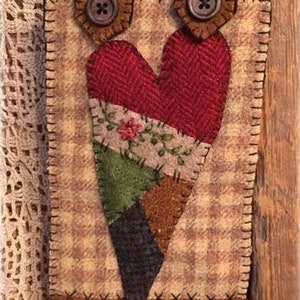 May include: A handmade, rustic-style wall hanging with a heart-shaped design. The heart is made of various fabric patches in red, green, brown, and black, with floral accents. It has brown leather straps with button closures and measures approximately 6 inches wide and 8 inches long.