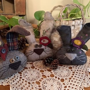 Może przedstawiać: Trzy ręcznie robione ozdoby zająca z filcu z haftowanymi detalami. Króliki są brązowe, szare i czarne z różnokolorowymi filcowymi kwiatami i guzikami na oczy.