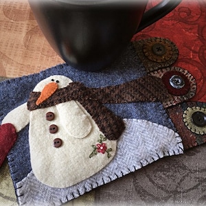 May include: A felt snowman with a brown scarf and a red heart in its hand. The snowman is on a blue and grey felt background with brown felt accents and buttons.