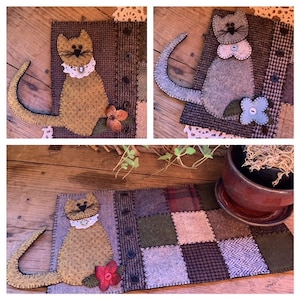 May include: A patchwork table runner with a felt cat appliqué. The cat is brown with a white collar and a red flower. The runner is made of various fabrics in shades of brown, green, and beige.