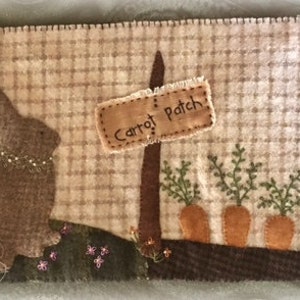 May include: A brown felt bunny sits near a sign that reads "Carrot Patch". The sign is attached to a brown stick. There are three green carrot plants with orange carrots growing in the ground. A small brown house with a red roof and a window is in the background.