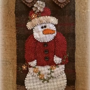 May include: A felt wall hanging featuring a snowman wearing a red jacket and a white hat with a brown button. The snowman is surrounded by green vines and yellow flowers. The hanging is brown with a plaid pattern and has brown buttons.